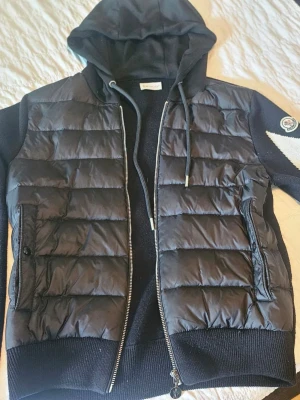 Svart dunväst med huva från Moncler - Snygg svart dunväst från Moncler med quiltad design och dragkedja framtill. Västen har en mjuk huva med dragsko och ribbade detaljer vid midjan. Klassisk Moncler-logga på ärmen och två praktiska fickor med dragkedja. Perfekt för lager-på-lager och streetstyle.