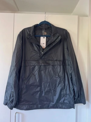 Svart vindjacka från NN07 - Snygg svart vindjacka från NN07 i storlek M. Jackan har halv dragkedja framtill, elastiska muddar och en minimalistisk design. Tillverkad i lätt, glansigt material som skyddar mot vind. Perfekt för dig som gillar stilrena och funktionella plagg.