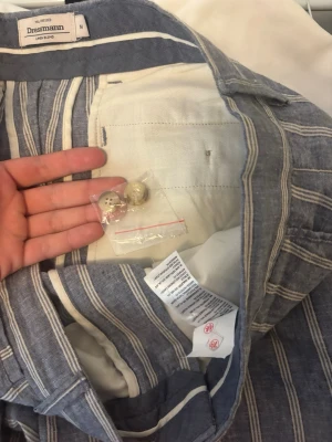 Blåvitrandiga linnebyxor Dressmann M - Snygga blåvitrandiga linnebyxor från Dressmann i storlek M. Byxorna är tillverkade i en linnemix och har klassiska vita vertikala ränder. Perfekta för en avslappnad och stilren look. Extra knappar medföljer. Aldrig använda