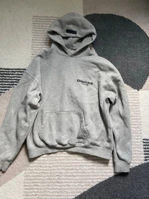 Grå hoodie Essentials Fear of God - Säljer en grå hoodie från Essentials Fear of God med svart logga på bröstet. Hoodien har en stor huva, magficka och ribbade muddar vid ärmar och nederkant. Tillverkad i mjukt bomullsmaterial som är perfekt för en avslappnad stil.