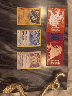 Pokémonkort Shining Rayquaza, Lugia och Arceus - Tre Pokémonkort: Shining Rayquaza, Shining Lugia och Shining Arceus. Alla kort har gul ram och holografisk yta. Medföljer tre Pokémon-kortfodral i rött, orange och lila med drakmotiv. Perfekt för samlare eller Pokémonfans.