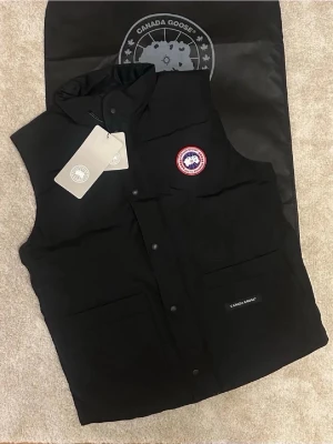 Svart dunväst från Canada Goose - Snygg svart dunväst från Canada Goose med klassisk logotyp på bröstet och två stora fickor framtill. Västen har hög krage, tryckknappar och är fylld med dun och fjädrar för extra värme. Perfekt för lager-på-lager och streetwear-stil.