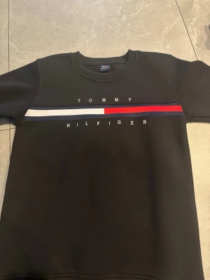 Svart sweatshirt Tommy Hilfiger - Snygg svart sweatshirt från Tommy Hilfiger med klassisk logga och broderad text på bröstet. Tröjan har rund halsringning och en rand i vitt, rött och marinblått över bröstet. Tillverkad i mjukt bomullsmaterial som är skönt att bära.