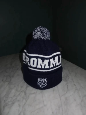 Marinblå Bromma mössa med tofs - Säljer en marinblå och vit mössa med stor text 'BROMMA' runtom och en broderad björnlogga framtill. Mössan har en klassisk tofs på toppen i matchande färger och är stickad i mjukt material. Perfekt för dig som vill representera Bromma med stil. Vet ej vilket märke