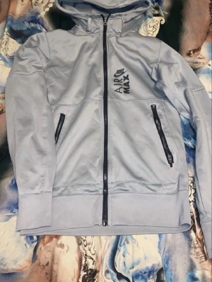 Ljusgrå Nike Air Max tracksuit - Snygg ljusgrå hoodie från Nike Air Max med dragkedja framtill och två svarta dragkedjeförsedda fickor. Nike Air Max-logga på bröstet. Tillverkad i mjukt material och har ribbade muddar vid ärmslut och nederkant. Perfekt för en sportig och avslappnad stil.