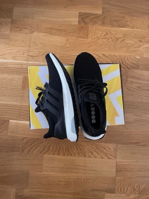 Adidas Ultra Boost svarta sneakers - Svarta Adidas Ultra Boost sneakers med stickad ovandel och vita Boost-sulor. Skorna har svarta plastdetaljer vid snörningen och hälpartiet samt en svart yttersula. Perfekta för dig som gillar sportig och stilren design.