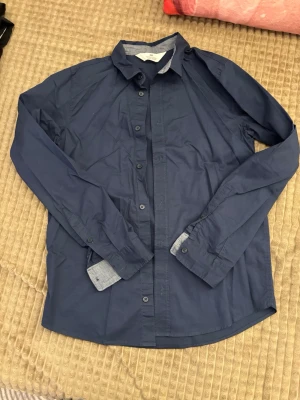 Mörkblå skjorta från H&M - Snygg mörkblå skjorta från H&M med klassisk krage och knappar framtill. Tillverkad i mjuk bomull och har långa ärmar med knapp vid manschetten. Perfekt att styla till jeans eller chinos för en clean look.