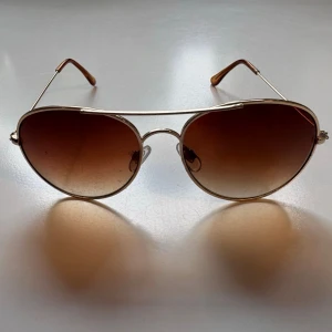 Guldiga aviator solglasögon - Snygga aviator solglasögon med guldfärgad metallbåge och bruna tonade glas. Klassisk design med dubbla bryggor och tunna skalmar. Perfekta för att lyfta din stil under soliga dagar.
