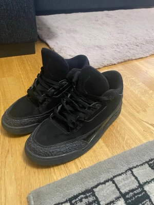 Svarta Air Jordan 3 black cats - Säljer ett par helsvarta Air Jordan 3 sneakers med klassisk siluett och detaljer i mocka och läder. Skorna har svarta skosnören, ikoniska elefantmönstrade paneler och Jumpman-logga på plösen. Perfekta för dig som gillar stilrena och sportiga sneakers.