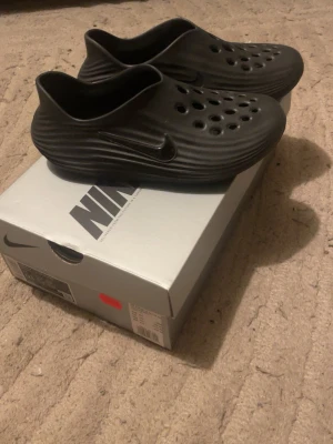 Svarta Nike tofflor med hålmönster - Säljer ett par svarta tofflor från Nike med vågig struktur och flera runda hål på ovansidan för ventilation. Tofflorna har en låg slip-on design och är tillverkade i ett lätt, formgjutet material. Perfekta för chill dagar eller att glida runt hemma.