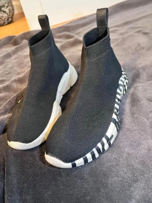 Love Moschino skor - Svarta sock-sneakers från Moschino med stretchig stickad ovandel och vit sula med svart textprint. Skorna har en chunky sula och dragflik bak för enkel påtagning. Perfekt för dig som gillar streetstyle och vill sticka ut med cool design. Priset kan diskuteras. SKORNA LUKTAR EJ.