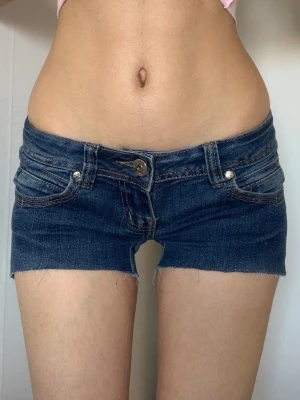 Lågmidjade jeansshorts i blå denim - Säljer ett par riktigt snygga lågmidjade jeansshorts i mörkblå denim. Shortsen har råa kanter, klassiska fickor fram och bak samt bälteshällor. Perfekta för varma dagar och ger en cool, avslappnad vibe.