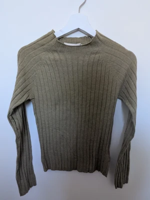 Olivgrön ribbstickad tröja - Säljer en olivgrön ribbstickad tröja med lång ärm och högre rund halsringning. Tröjan har en smal passform och är tillverkad i ett mjukt, stretchigt material. Perfekt för lager på lager och enkel att matcha med olika outfits. Inte används alls och finns någon sorts av lapp kvar men det är inte en prislapp. Skriv gärna om ni har några frågor💕