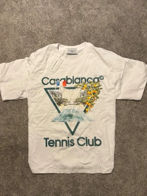 Vit Casablanca Tennis Club t-shirt - Snygg vit t-shirt från Casablanca med tryck framtill som visar ett tennisinspirerat motiv och texten 'Casablanca Tennis Club'. T-shirten har rund hals och korta ärmar. Perfekt för dig som gillar sportig och trendig stil. Storlek s men passar m