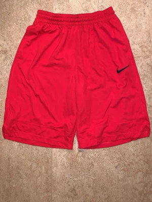 Röda Dri-FIT shorts från Nike - Säljer ett par röda sportiga shorts från Nike med elastisk midja och svart Nike-logga på benet. Tillverkade i lätt och snabbtorkande Dri-FIT material som passar perfekt för träning eller varma dagar.