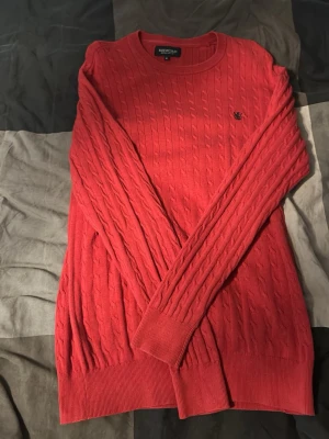 Röd kabelstickad tröja Redford - Snygg röd kabelstickad tröja från Redford med rund halsringning och diskret logga på bröstet. Tröjan har långa ärmar och ribbade muddar vid ärmslut och nederkant. Perfekt för dig som gillar klassisk stil med en modern touch.