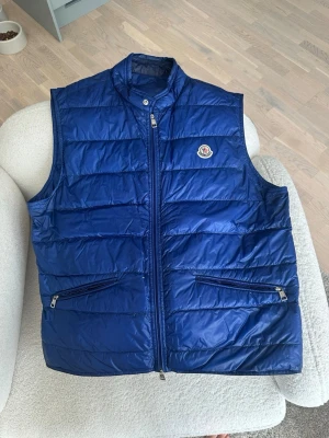 Moncler GUI väst - Snygg blå dunväst från Moncler med quiltad design och hög krage. Västen har två dragkedjeförsedda fickor framtill och en liten Moncler-logga på bröstet. Perfekt att slänga över en hoodie eller tröja för extra värme och stil.