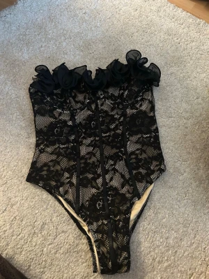Svart spetsbody med volangdetalj - Snygg svart body i spets med blommönster och volangdetalj upptill. Axelbandslös modell med beige foder under spetsen som ger en lyxig känsla. Perfekt för dig som vill ha något extra till din outfit. Jag har provat den men inte använt den!