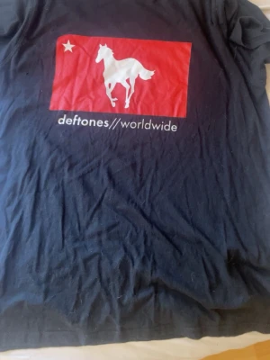 Deftones svart band-tshirt - Svart t-shirt från Deftones med ikoniskt tryck av en vit häst på röd bakgrund och texten 'deftones // worldwide' framtill. Köpt för 700kr från Redrum 