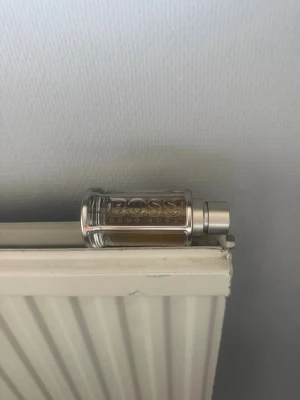 Hugo Boss BOSS parfymflaska - Snygg parfymflaska från Hugo Boss med genomskinligt glas och silverfärgad topp. Flaskan har en stilren, cylindrisk form och texten BOSS ingraverad på framsidan. Innehållet är ljusbrunt och flaskan känns lyxig och modern.