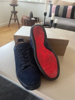 Mörkblå sneakers från Christian Louboutin - Snygga mörkblå sneakers i mocka från Christian Louboutin med klassisk röd sula. Skorna har svarta skosnören, låg profil och diskreta sömmar. Perfekta för dig som vill sticka ut med exklusiva detaljer och lyxig känsla.