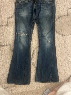 Blå bootcut jeans från True Religion - Säljer ett par blå bootcut jeans från True Religion med slitningar och hål på framsidan. Jeansen har dekorativa bakfickor med kontrastsömmar och knappar. Klassisk femficksmodell i denim med cool vintagekänsla.