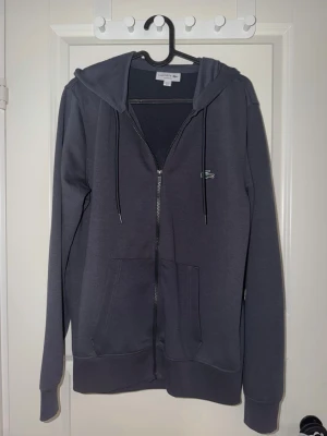 Mörkgrå zip-hoodie från Lacoste - Snygg mörkgrå hoodie från Lacoste med dragkedja framtill och klassisk krokodillogga på bröstet. Tröjan har huva med dragsko och två stora fickor framtill. Perfekt för en chill och stilren look.