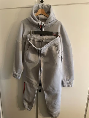 Grått loungewear-set från Prada - Snyggt grått loungewear-set från Prada med hoodie och matchande byxor. Hoodien har dragkedja, stora fickor och röd logodetalj. Byxorna har resår i midjan och benslut samt dragsko. Perfekt för chill dagar hemma eller när du vill vara bekväm men ändå stilren.