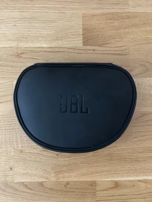 JBL over-ear hörlurar med fodral - Säljer ett par svarta over-ear hörlurar från JBL med tillhörande hårt fodral. Hörlurarna har rund form och vadderade kåpor med blå insida. Perfekta för musik, gaming eller film. Sladd medföljer och allt får plats i det smidiga fodralet.