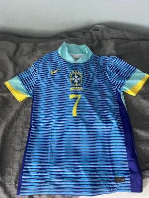 Blå Brasilien fotbollströja Nike - Säljer en blå och ljusblå randig Brasilien fotbollströja från Nike med gula detaljer och nummer 7. Tröjan har VINI JR. tryckt på ryggen och Brasil-logga på bröstet. Tillverkad i lätt och ventilerande material, går att sänka pris vid snabb affär 