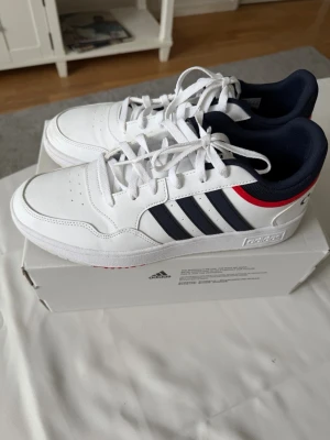 Vita Adidas sneakers med svarta ränder - Snygga vita sneakers från Adidas med klassiska svarta ränder på sidorna och en röd detalj vid hälen. Skorna har lågt skaft, vit sula och mörkblå insida. Perfekta för en sportig och stilren look. Tillverkade i syntetmaterial och textil.