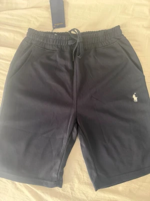 Svarta shorts från Polo Ralph Lauren - Snygga svarta shorts från Polo Ralph Lauren med klassisk logga broderad på benet. Shortsen har dragsko i midjan, två sidofickor och är tillverkade i mjuk bomull. Perfekta för en avslappnad och stilren look.