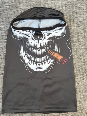 Balaclava med dödskalle och cigarr - Svart balaclava i stretchigt syntetmaterial med ett stort vitt dödskalletryck och en brun cigarr med röd glöd. Masken har öppning för ögonen och täcker hela huvudet. Perfekt för dig som vill ha en unik och edgy accessoar.