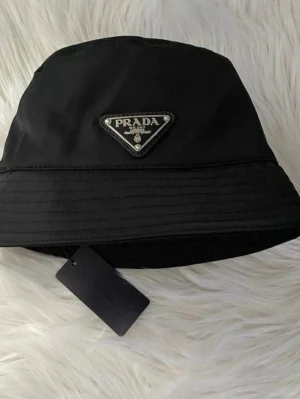 Svart bucket hat från Prada - Stilren svart bucket hat från Prada med klassisk trekantig logotyp framtill. Hatten är tillverkad i ett glansigt nylonmaterial som ger en modern vibe. Perfekt accessoar för att lyfta din streetwear-look.