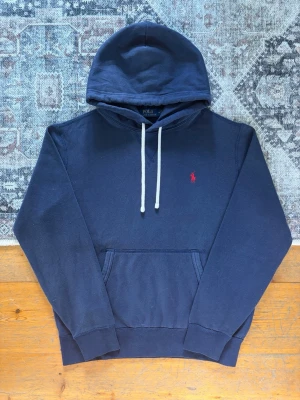 Marinblå Polo Ralph Lauren Hoodie - Storlek M - Hej! 👋 Säljer en klassisk marinblå Polo Ralph Lauren hoodie i storlek M med röd pony-logga. 🐎✨ Tröjan är i gott skick överlag men har blivit något nopprig. Det finns även en mindre, nästintill osynlig fläck på magfickan (se sista bilden). 100% äkta med originaltvättlappar kvar och en fullt fungerande QR-kod för superenkel verifiering ✅ Ett klockrent och tidlöst premium-basplagg! Fraktar snabbt! 🚛📦💬 