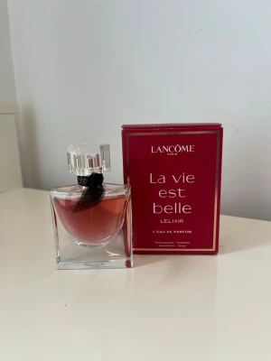 Lancôme La Vie Est Belle L'Elixir parfym - Lancôme La Vie Est Belle L'Elixir är en elegant parfym i en fyrkantig glasflaska med rosaröd vätska och en dekorativ svart rosett vid halsen. Kommer med en snygg röd kartong med guldiga detaljer. Perfekt för dig som gillar lyxiga dofter. Använd några gånger, nästan full.