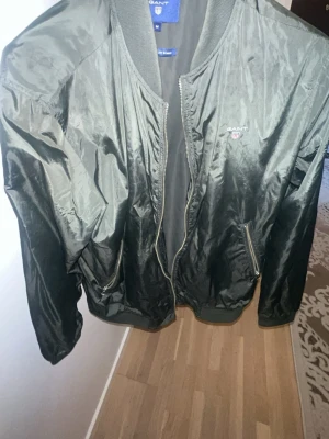 Svart bomberjacka från Gant - Snygg svart bomberjacka från Gant i nylon med klassisk passform. Jackan har dragkedja framtill, två fickor med dragkedja och ribbade muddar vid ärmslut och krage. Gant-logga broderad på bröstet. Perfekt för en stilren look.
