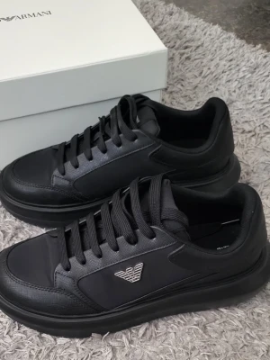 Emporio armani sneakers - Stilrena svarta sneakers från Armani med diskret logga i silver på sidan. skosnören och är tillverkade i en mix av läder och textil. Perfekta för dig som gillar en clean och modern look.