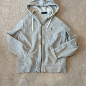 Ljusgrå hoodie från Polo Ralph Lauren - Snygg ljusgrå hoodie från Polo Ralph Lauren med dragkedja, huva med snörning och broderad logga på bröstet. Mjuk insida och två fickor framtill. Perfekt för chill dagar eller när du vill ha en clean och stilren look. Lite varmare material som håller dig varm i lite kyligare väder. Denna modell säljs inte längre av Ralph Lauren därför är den ”sällsynt”