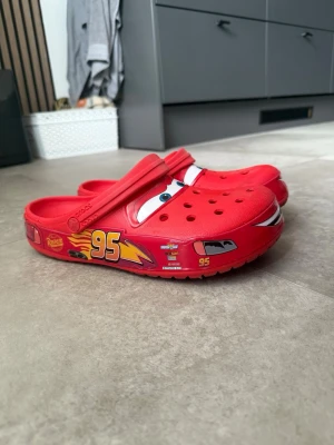 Röda Crocs Cars Lightning McQueen - Säljer ett par skojiga röda Crocs-tofflor med Lightning McQueen-motiv. Tofflorna har detaljer som ögon, nummer 95 och blixtar på sidorna. Tillverkade i lätt och tålig plast med justerbar hälrem för extra komfort.