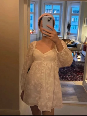 Vit kort klänning med spetsdetaljer - Söt vit kortklänning med v-ringning och långa puffärmar. Klänningen har fina spetsdetaljer och knappar framtill, samt ett romantiskt mönster i tyget. Perfekt för sommarens alla tillfällen!