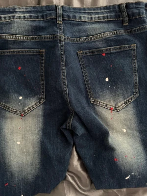 Mörkblå jeans med patch och färgstänk - Säljer ett par mörkblå jeans med vit och röd färgstänk samt en stor svartvit patch på vänster sida fram. Jeansen har slitningar, fem fickor och kontrastsömmar. Perfekt för dig som gillar en unik och streetinspirerad stil.