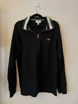Lacoste halv-zip - Snygg svart långärmad ziptröja från Lacoste med klassisk grön logga på bröstet. Tröjan har en halv dragkedja och en krage med vita och gröna detaljer. Perfekt för en sportig och stilren look.