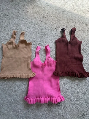 Tre ribbade linnetoppar med volang - Tre snygga ribbade linnetoppar i beige, rosa och brun. Alla har breda axelband, v-ringning och volangkant nertill. Perfekta att styla med jeans eller kjol för en trendig look. Säljer dem styck (89kr/st)!!🫶