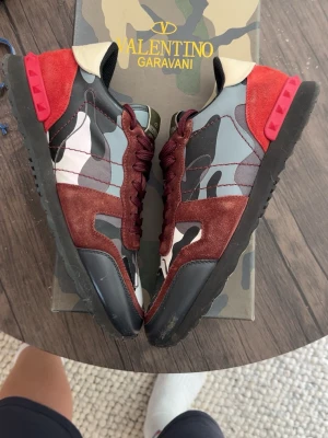 Valentino Garavani rockrunners  - Snygga sneakers från Valentino Garavani med kamouflagemönster i grått, svart och vitt, detaljer i vinrött mocka och röd häl. Skorna har svarta sulor och olivgröna plösar med logga. Perfekta för dig som vill sticka ut med exklusiv streetstyle.