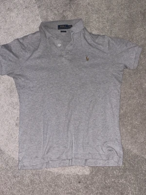 Grå pikétröja från Polo Ralph Lauren - Klassisk grå pikétröja från Polo Ralph Lauren med liten broderad logga på bröstet. Tröjan har korta ärmar, krage och knappar framtill. Perfekt basic till garderoben och enkel att matcha. Perfekt för sommar väder.