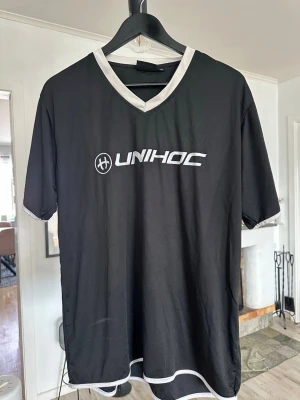 Svart Unihoc sporttröja med vit kant - Säljer en svart sporttröja från Unihoc med vit V-ringad krage och vita detaljer vid ärmslut och nederkant. Tröjan har korta ärmar och är tillverkad i ett lätt och funktionellt material, perfekt för innebandy eller annan träning.