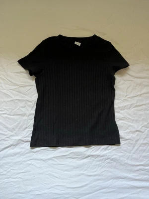 Svart ribbad t-shirt från Lindex - En svart ribbad t-shirt från Lindex med rund hals och korta ärmar. Tillverkad i mjuk bomull och har en enkel, stilren look som passar till allt. Perfekt basplagg för dig som gillar minimalistisk stil.