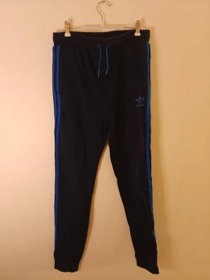 Adidas mjukisbyxor  - Säljer ett par mörkblå mjukisbyxor från Adidas med klassiska blå ränder längs sidorna och dragsko i midjan. Byxorna har smal passform och Adidas-logga på vänster ben. Perfekta för chill eller träning. Storlek XL för barn.