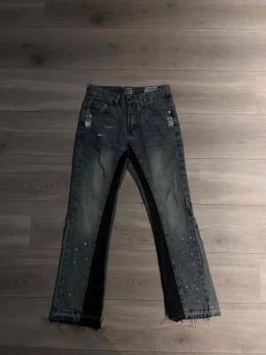Mörkblå svart gallery dept. - Unika grå jeans med svarta paneler längs insidan av benen och råa, fransiga benslut. Jeansen har slitna detaljer, vita färgstänk och klassisk femficksmodell. Perfekt för dig som vill sticka ut med en edgy streetwear-look.